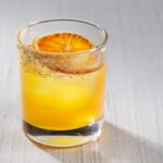 An orange cocktail in a short glass with an orange slice inside all on a white table.