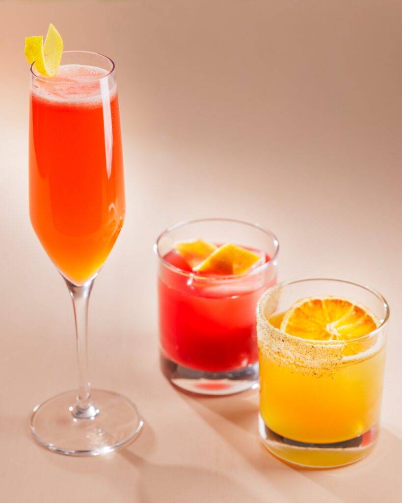 A tall redish cocktail with a lemon twist beside a short red cocktail and an orange cocktail with an orange slice on top. 