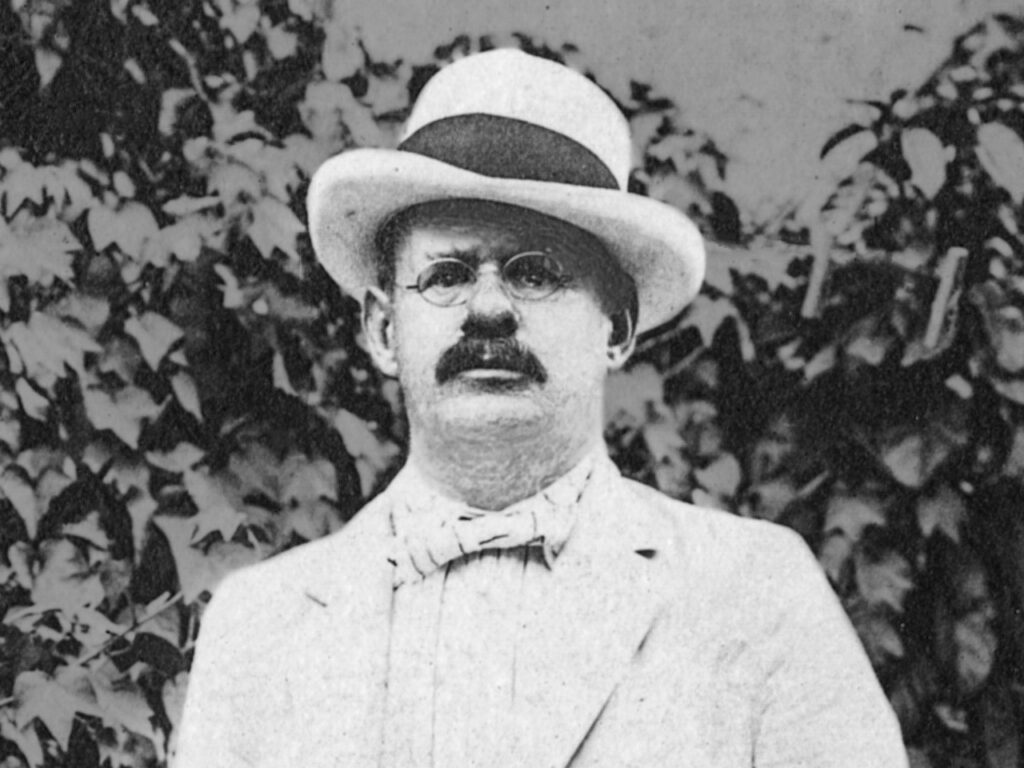 A man in a black and white photo with a hat, mustache, and glasses.