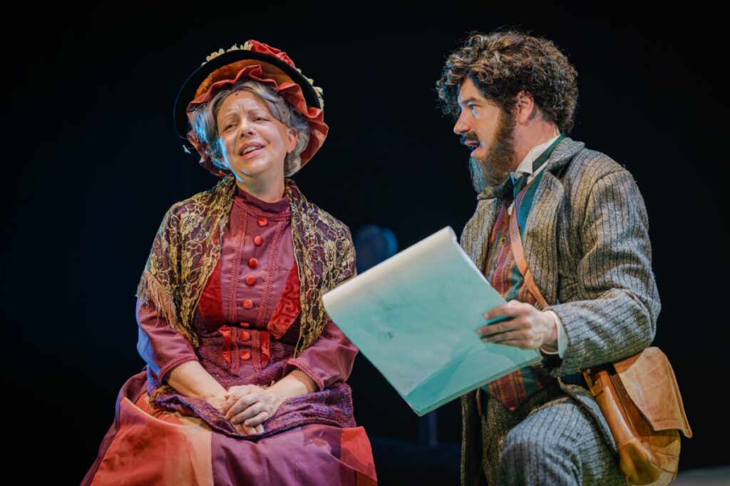 Live musical theater performance showing an actor with a sketchpad and an actress in a vintage plum dress singing.
