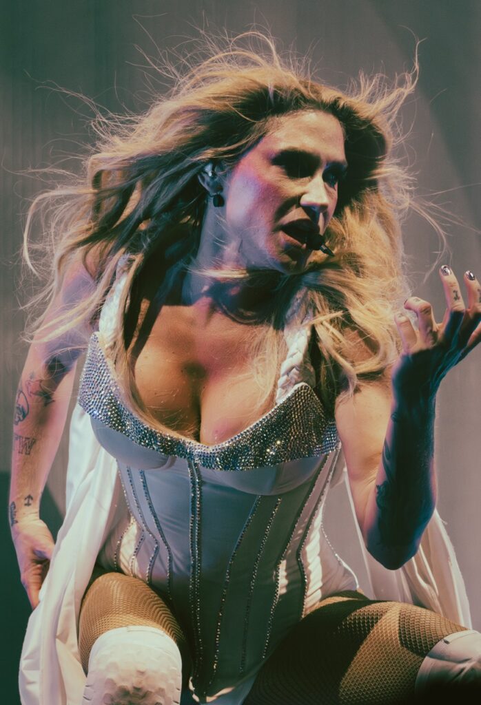 Kesha in a white corset top kneeling down on stage with her hair blown back.