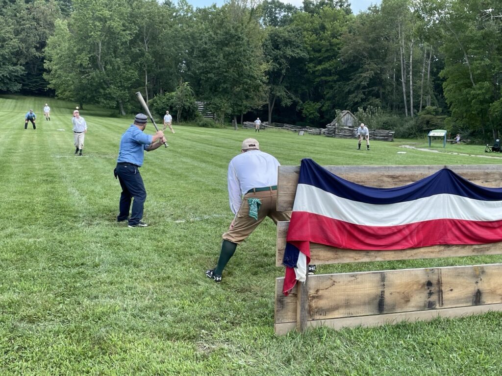 Vintage base ball players on the field with a batter, catcher, pitcher, and other players in the field.