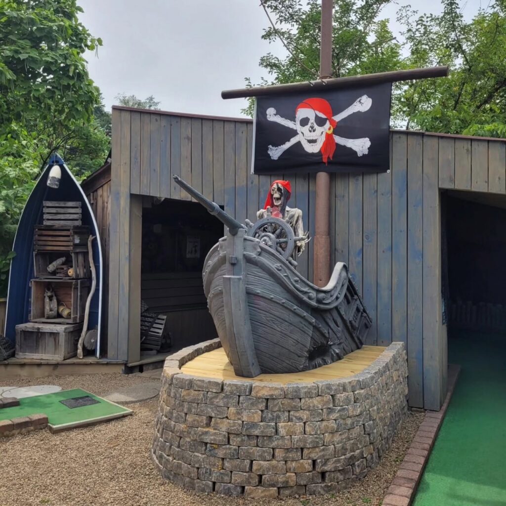 A pirate ship in front of a shed on a miniature golf course.