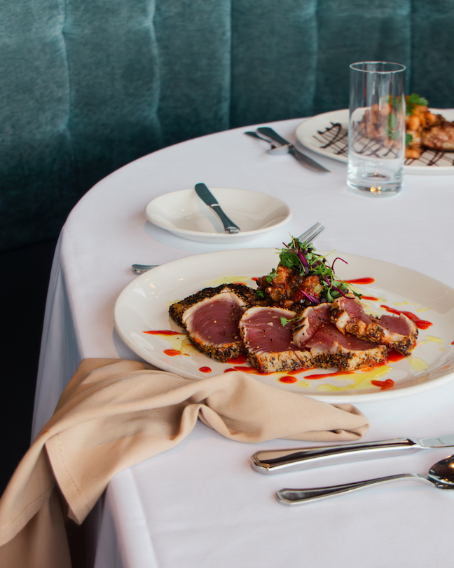 A plate of seared tuna on a table covered with a white linen in front of a green velvet booth.