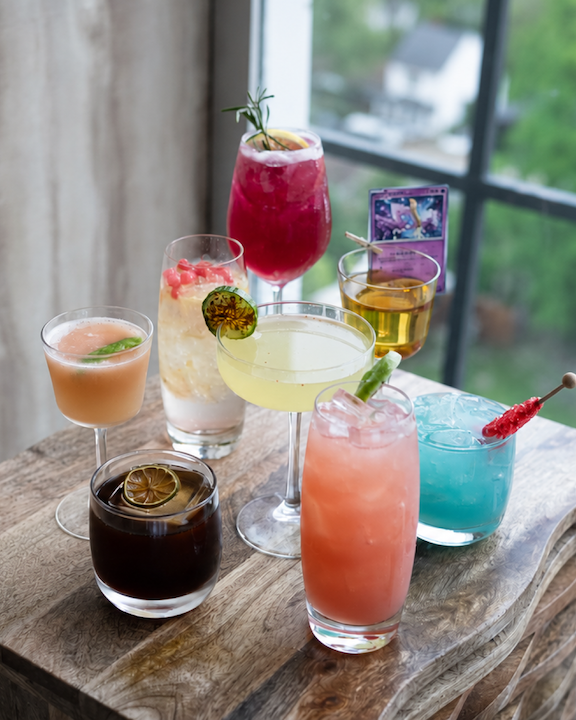 A variety of cocktails on a wooden table in front of a window.