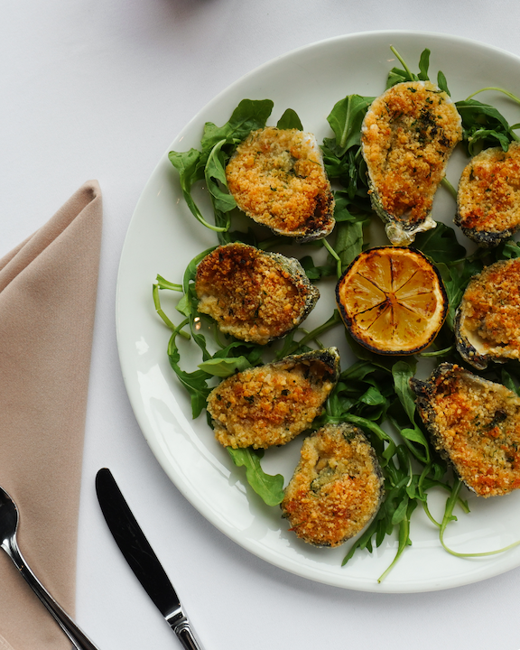 Oysters Rockefeller plated on arugula on a white plate with a grilled lemon in the center.
