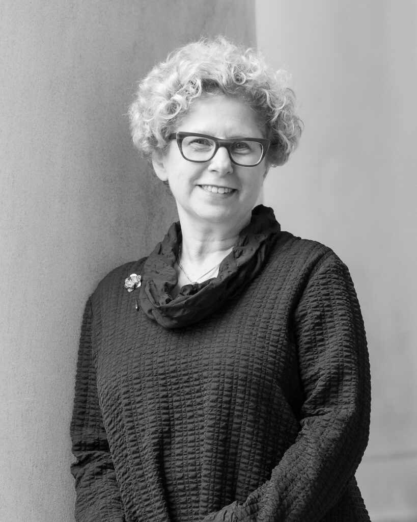 Nancy Zionts in a sweater and glasses standing in the corner of two walls. 