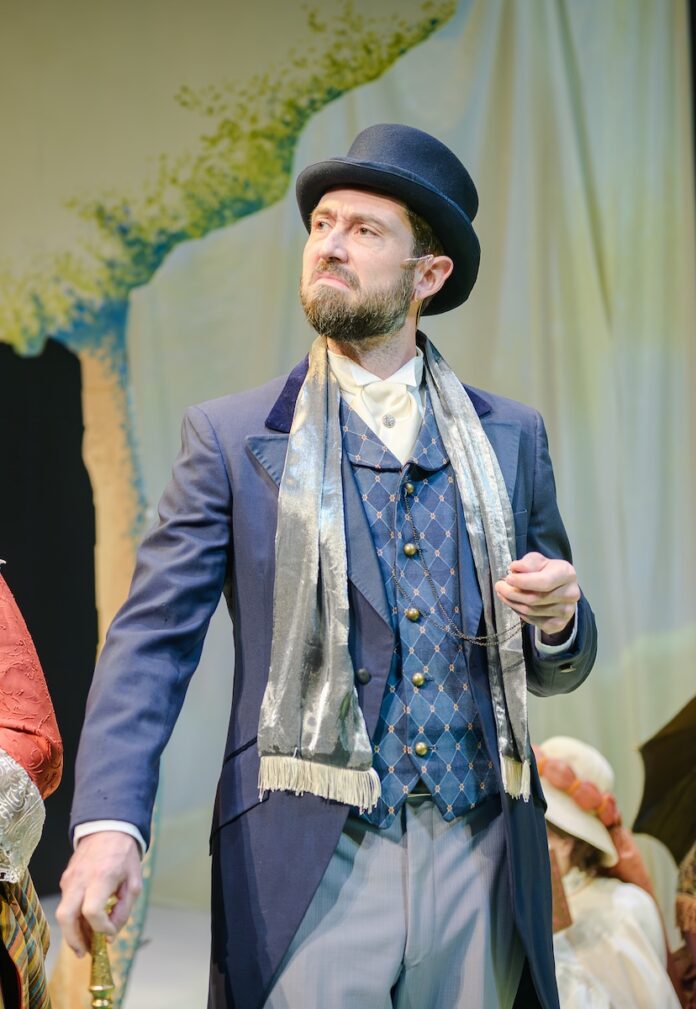 A man in a musical performance on stage in highbrow blue clothing with a cane and a hat, holding his pocketwatch in his hand.