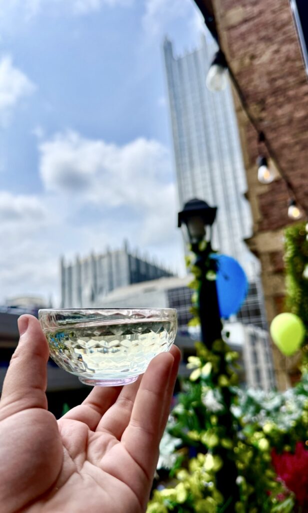 A perosn holds a small glass of Sake in front of the Pittsburgh cityscape Downtown.