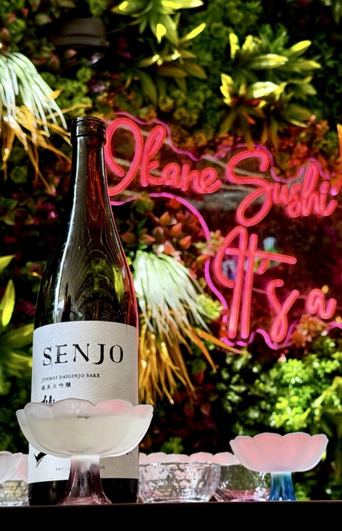 Okane Downtown Japanese Restaurant TABLE Magazine 11 A bottle of sake in front of a greenery wall and Okane Sushi sign.