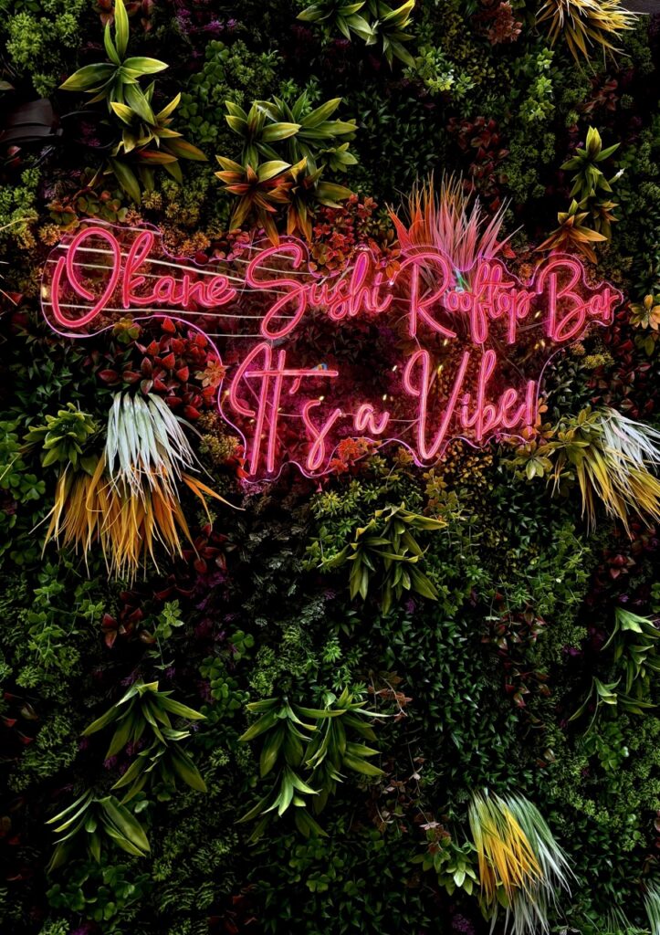 A green flower wall with a neon sign that reads "Okane Sushi Rooftop Bar, It's a Vibe"