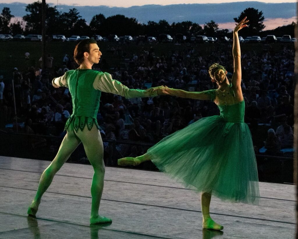Two ballet dancers in a green dress and green tights dance on stage as the man spins the woman out.
