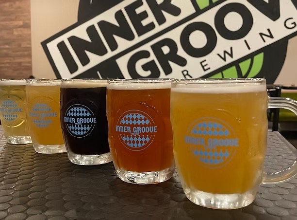 Pints of beer in various colors sit on a bar in front of the Inner Grove Brewing sign.