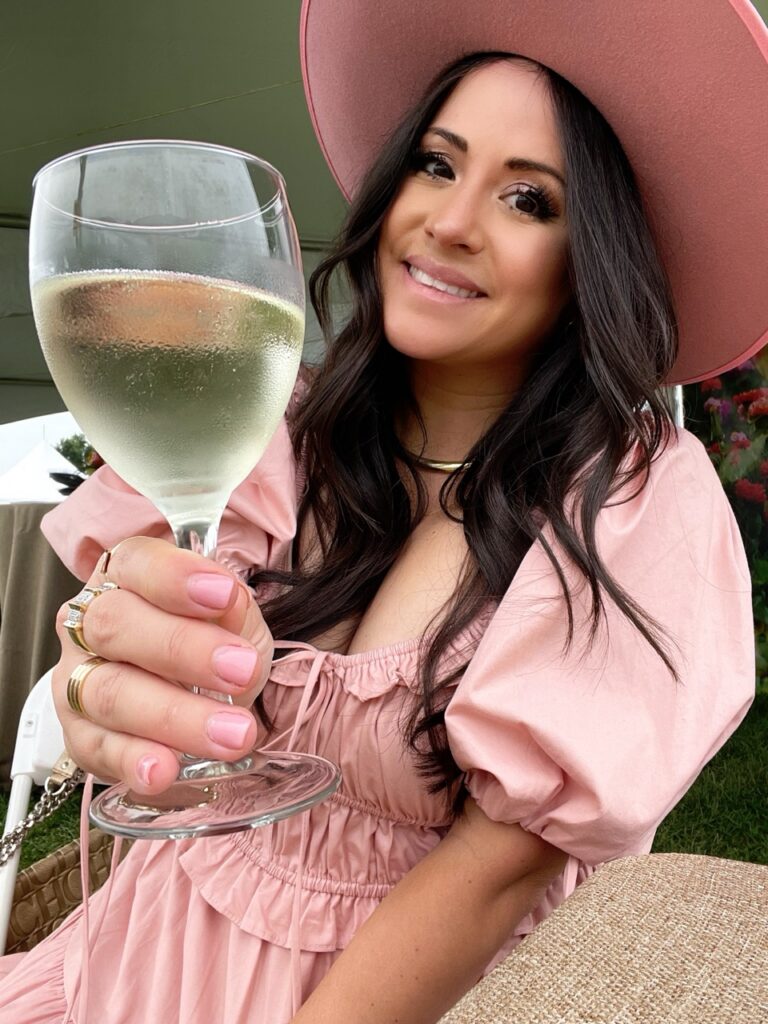 A woman in a hat reaching out her hand with a wine glass.