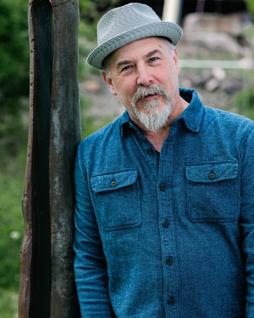 Actor Jeffrey Carpenter, playing Prospero in Quantum Theatre's The Tempest, leaning against a rusty metal beam.