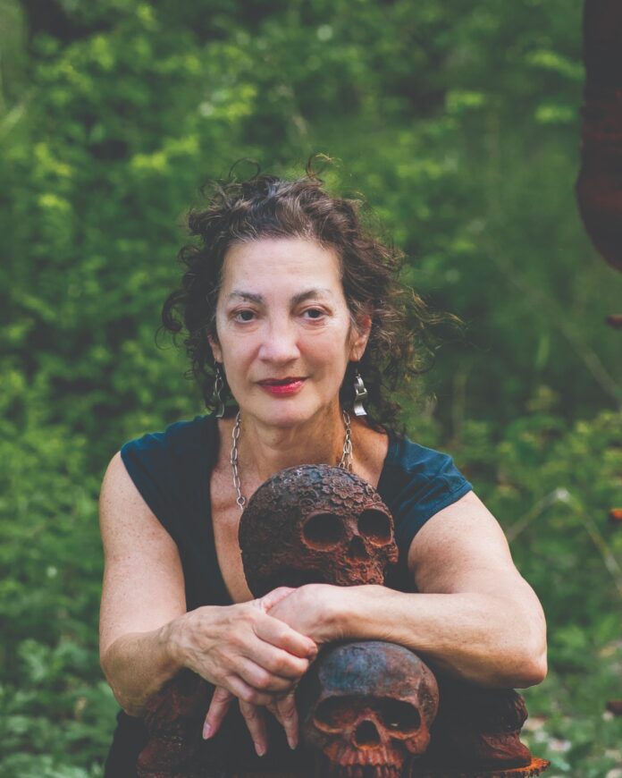 Karla Boos, retiring founder of Quantum Theatre, leaning on rusty metal skulls in a natural outdoor setting.