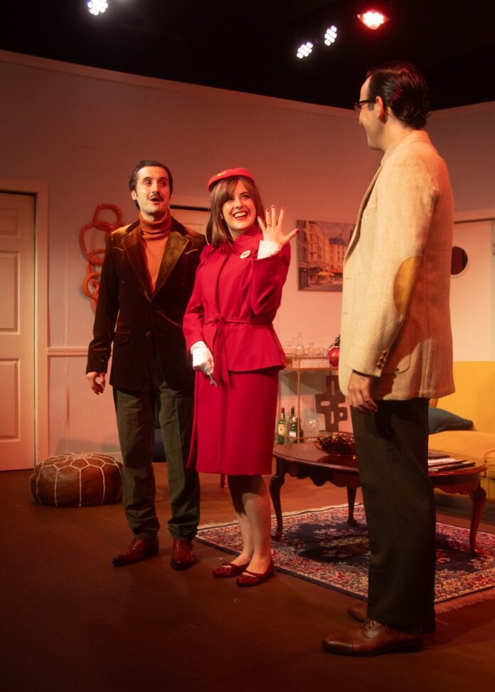 Two men in suit jackets and a woman in red clothing in a living room on stage.