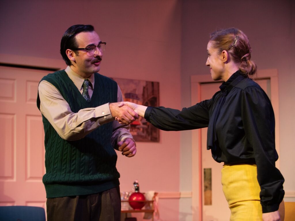 A woman in a black long sleeve shirt and yellow pants shakes hands with a man in a tie and sweater vest.