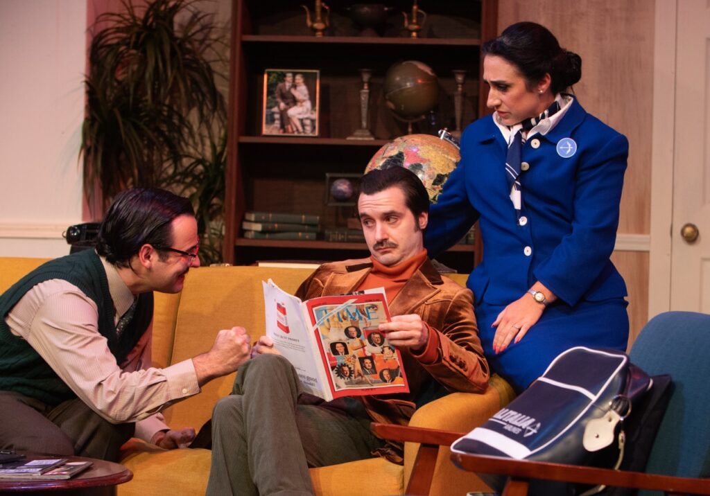 Two men sit on a couch while a woman in a blue airline costume sits on the arm of the couch.