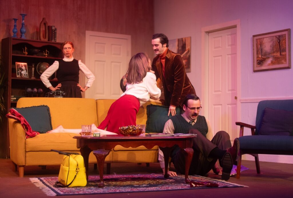 A woman leans on her knees on a yellow couch to a man as another man sits on the floor and a woman stands with her hands on her hips behind the couch.