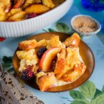 A serving of peach and blackberry bread pudding with golden bread chunks and cream on a brown plate over a floral cloth.