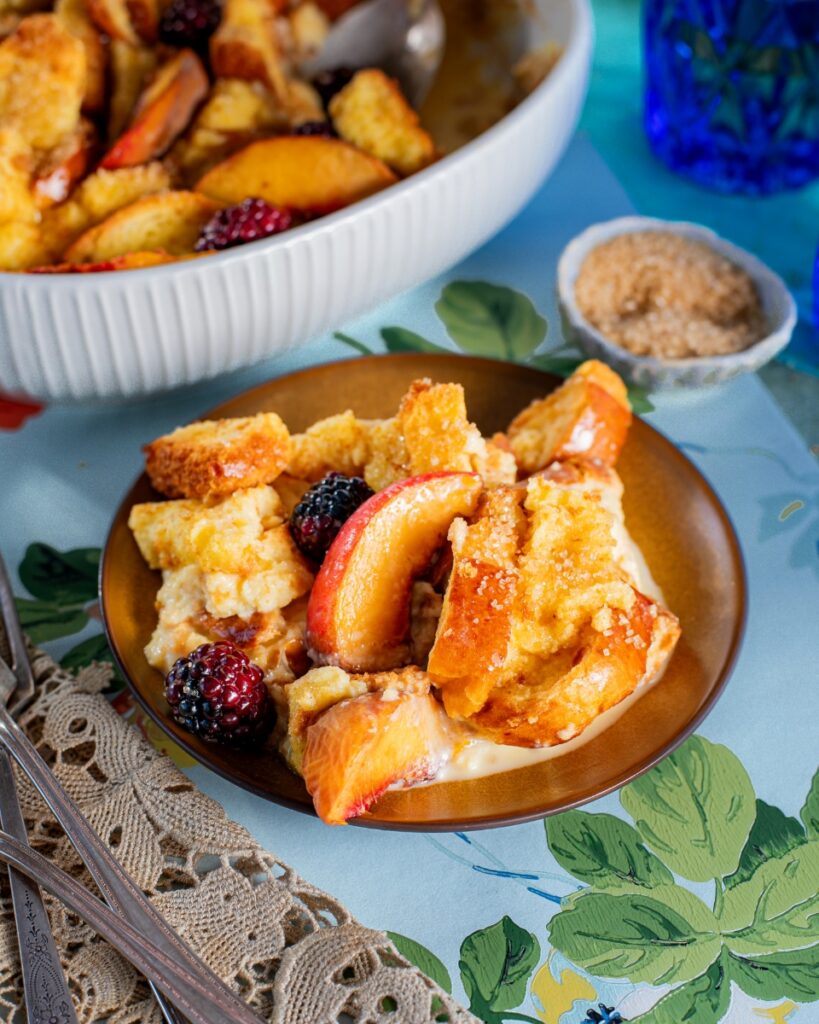 A serving of peach and blackberry bread pudding with golden bread chunks and cream on a brown plate over a floral cloth.
