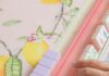The Magic of Mah Jongg and Why This Classic Game Is Making a Comeback Close-up of a player's hand moving pastel Mah Jongg tiles on a pink acrylic rack over a lemon-patterned mat.