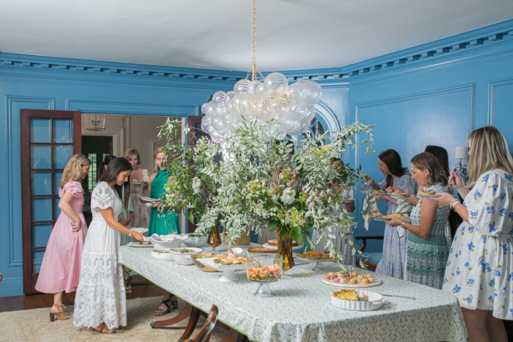 Guests serving themselves at a stylish Mah Jongg potluck luncheon in a blue dining room with an English garden floral centerpiece.