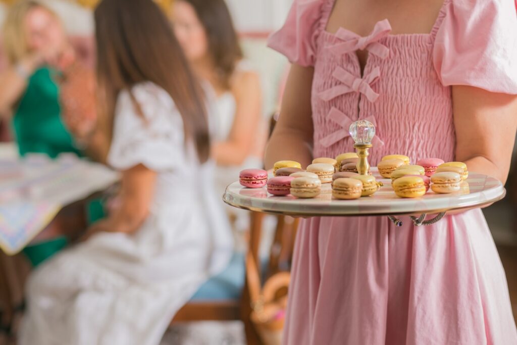 Woman in a pink summer dress holding a decorative tray of colorful macarons at a Mah Jongg gathering.