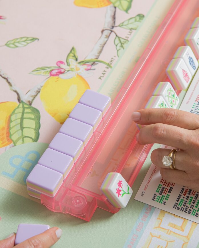 Close-up of a player's hand moving pastel Mah Jongg tiles on a pink acrylic rack over a lemon-patterned mat.