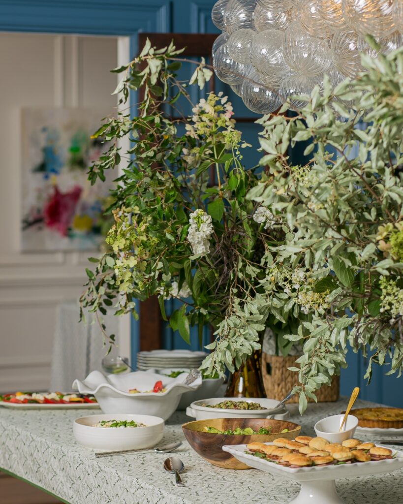 Potluck buffet table featuring light summer fare and a large, organic floral arrangement of hydrangeas and eucalyptus.