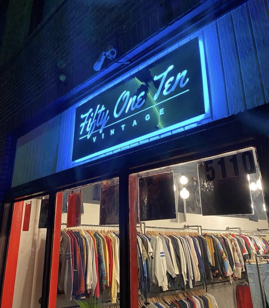 The outside of Fifty One Ten thrift store with a light up blue sign and racks of clothes in the window.