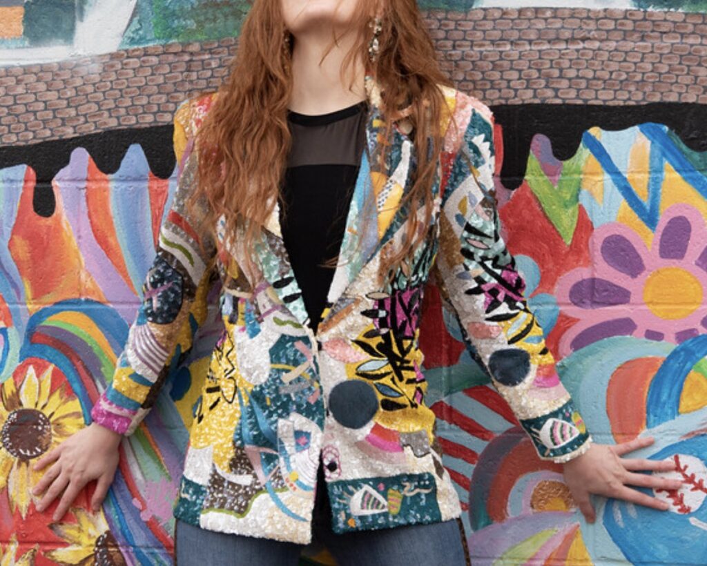 A person with long orange hair wears an abstract art blazer in front of an abstract colorful art piece.