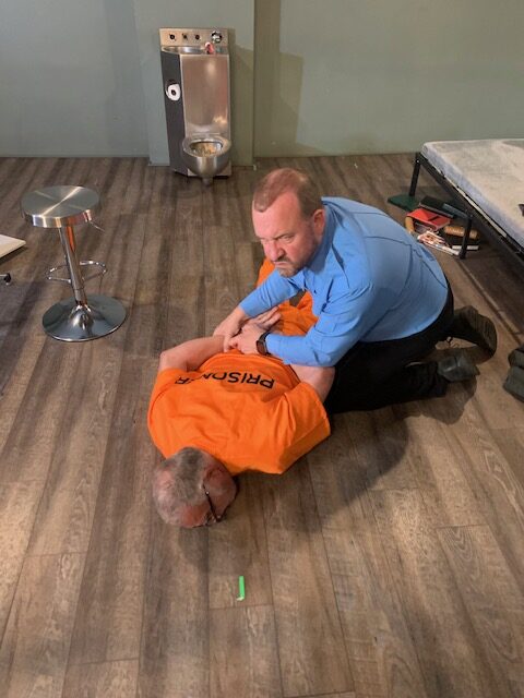 Man in blue shirt restraining prisoner in orange jumpsuit in a cell