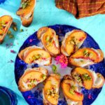 Whipped goat cheese and peach bruschetta sprinkled with pistachios on a blue patterned plate over a turquoise surface.