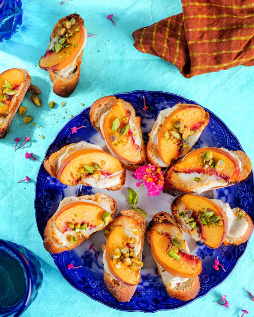 Whipped goat cheese and peach bruschetta sprinkled with pistachios on a blue patterned plate over a turquoise surface.