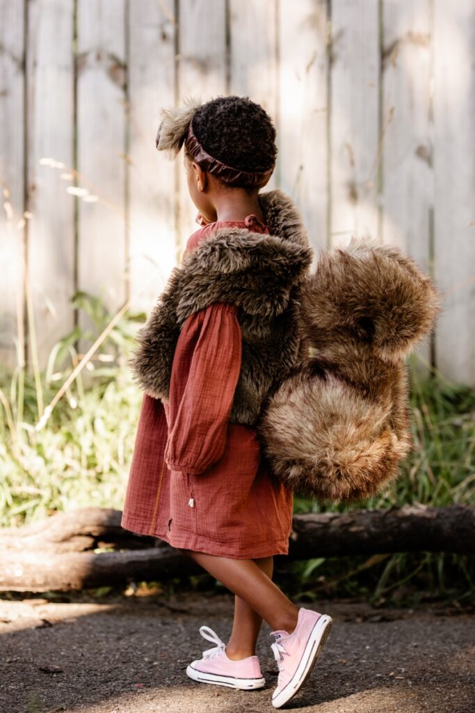 Profile shot of a child dressed in a squirrel costume featuring a plush faux-fur tail, a fur vest, and ears on a headband, wearing a rust-colored dress and pink sneakers.