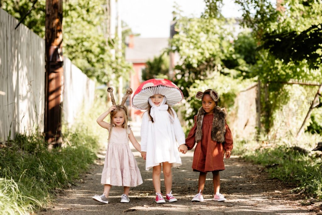 Three young children standing hand-in-hand outdoors in DIY costumes representing a snail, a red-and-white toadstool mushroom, and a squirrel.