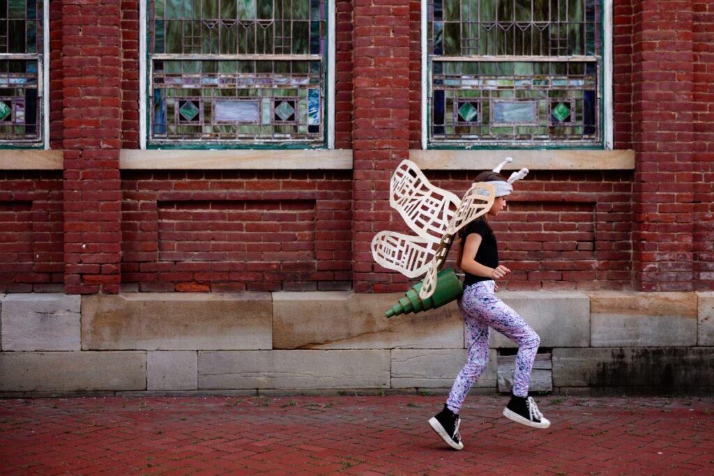 Side view of a child in a dragonfly costume wearing cutout cardboard wings, a green segment tail, patterned leggings, and antennae headband, walking along a brick wall.