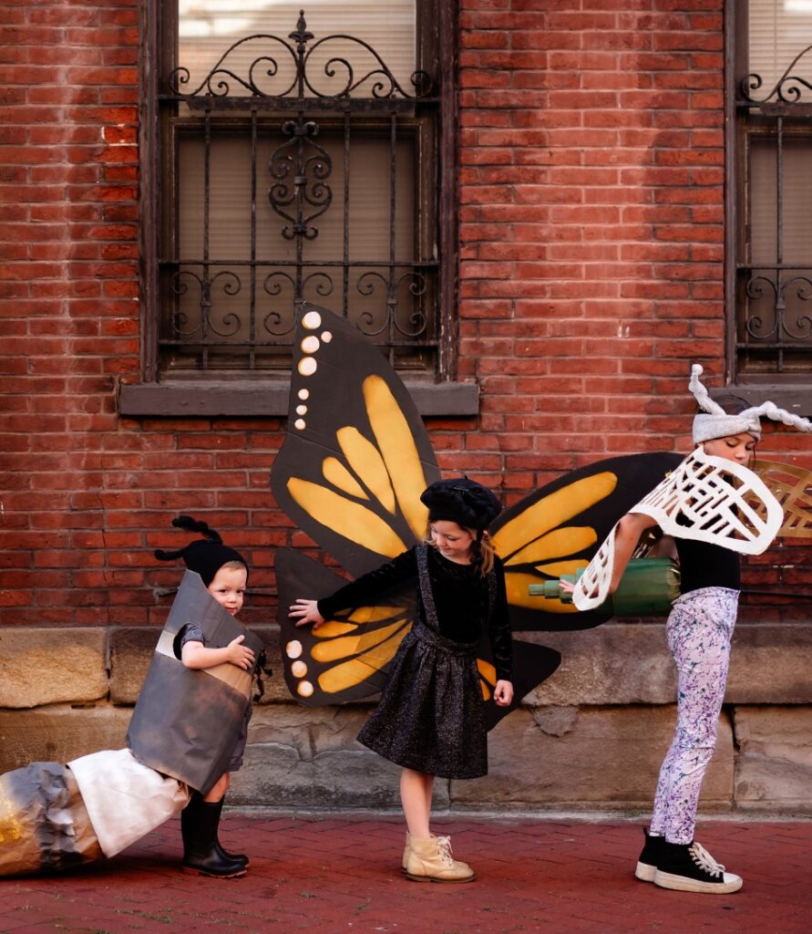 Three children outdoors in homemade nature costumes: a caterpillar on the left, a monarch butterfly in the center, and a dragonfly on the right, set against a red brick building.