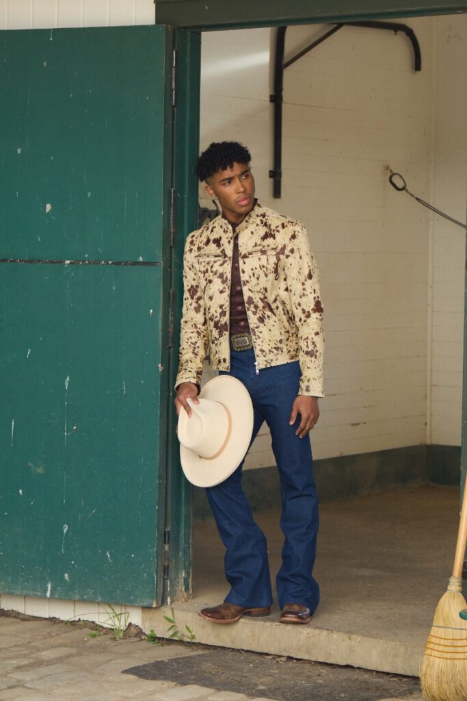 A young man in a cow-hide print jacket, dark jeans, cowboy boots, and holding a beige cowboy hat stands in a green barn doorway.