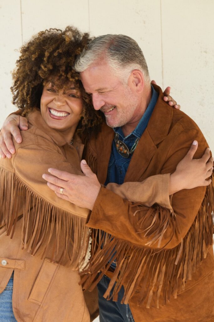 A smiling couple embracing while wearing matching brown suede jackets with fringe detail along the arms and shoulders.