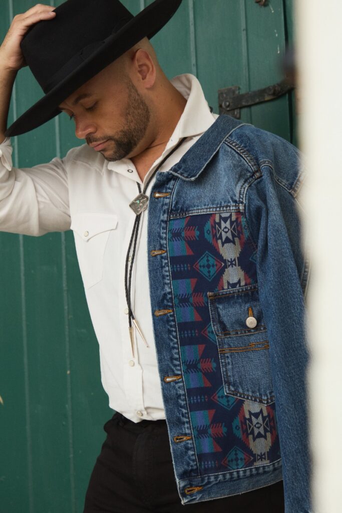 A close-up profile of a man wearing a black cowboy hat, a bolo tie, a white shirt, and a denim jacket with a patterned fabric panel.