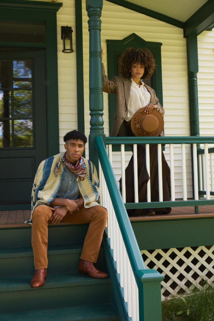 Two models posed on porch steps of a farm building: a man seated in a printed wrap and corduroys, and a woman above in a tweed blazer and hat.