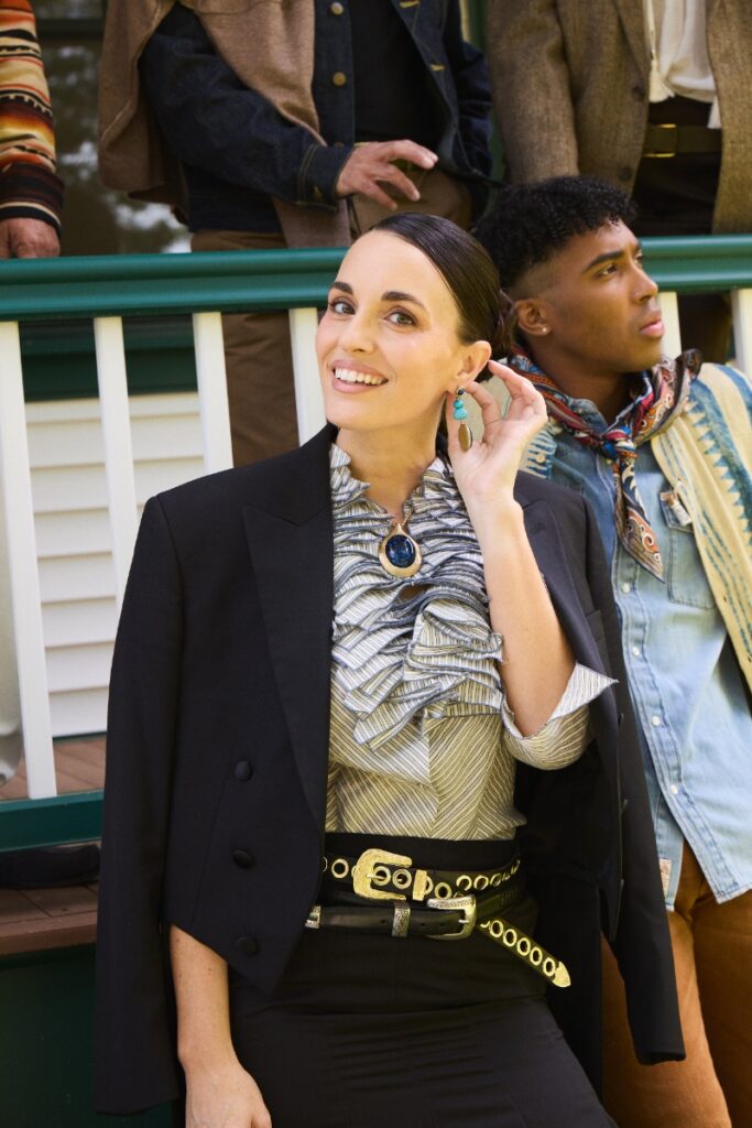 A woman smiling in a black blazer, ruffled blouse, statement pendant, and western belt, with another model seated behind her.