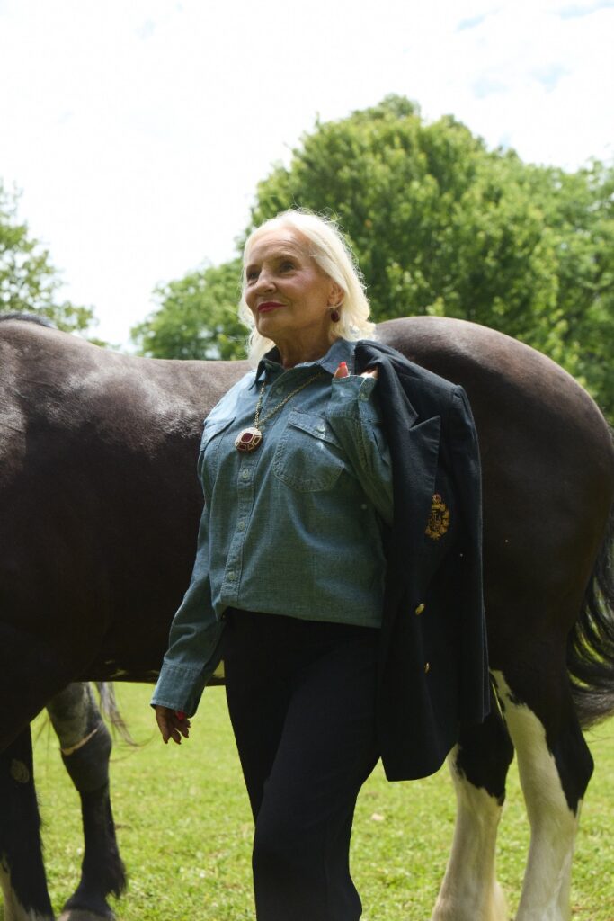 A blonde woman walking beside a dark horse outdoors, wearing a denim button-down shirt, black pants, and a dark blazer draped over her shoulder.