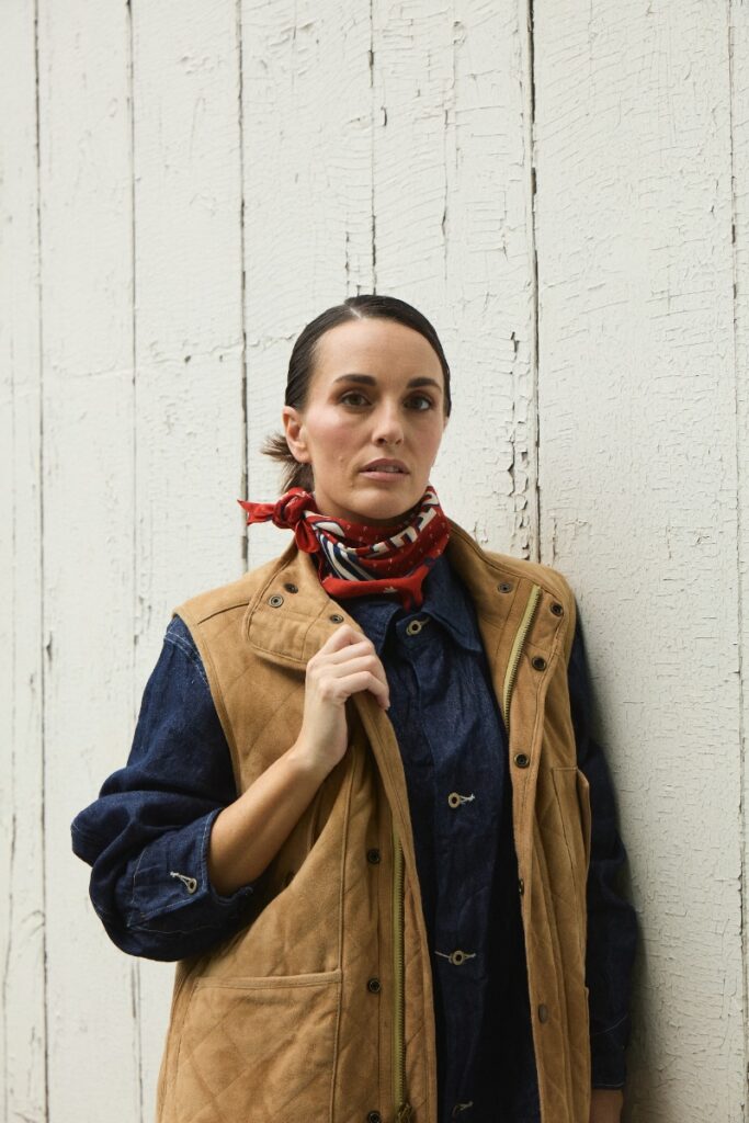 A woman leaning against a white weathered wooden wall wearing a quilted tan vest over a denim shirt and a red neck kerchief.