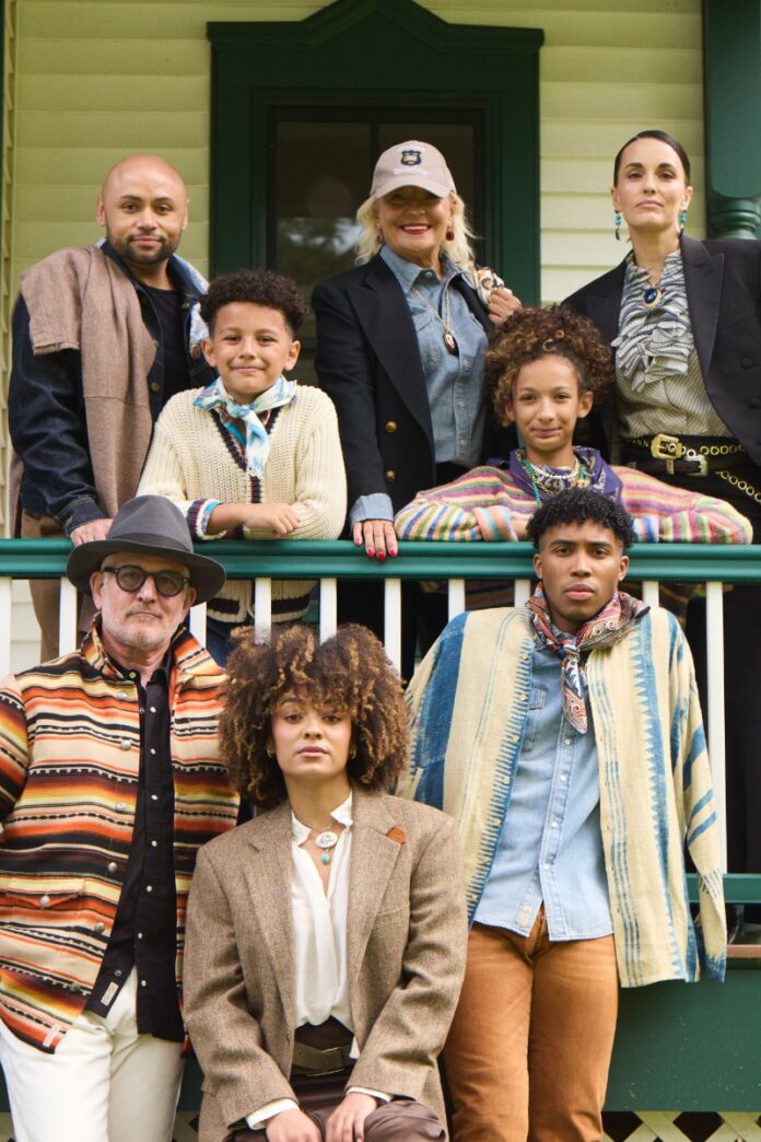 A group portrait of eight people of various ages wearing layered autumn fashion on a porch, styled with hats, blazers, and knits.