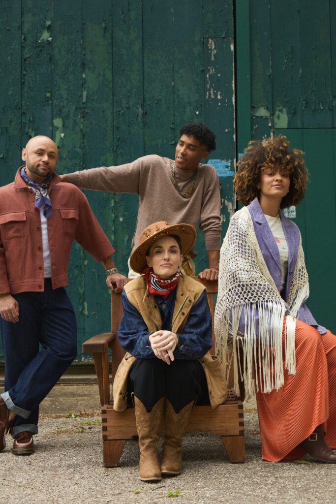 Four models posed in layered autumn attire outdoors in front of a green barn door, incorporating western hats, fringed shawls, and cowboy boots.