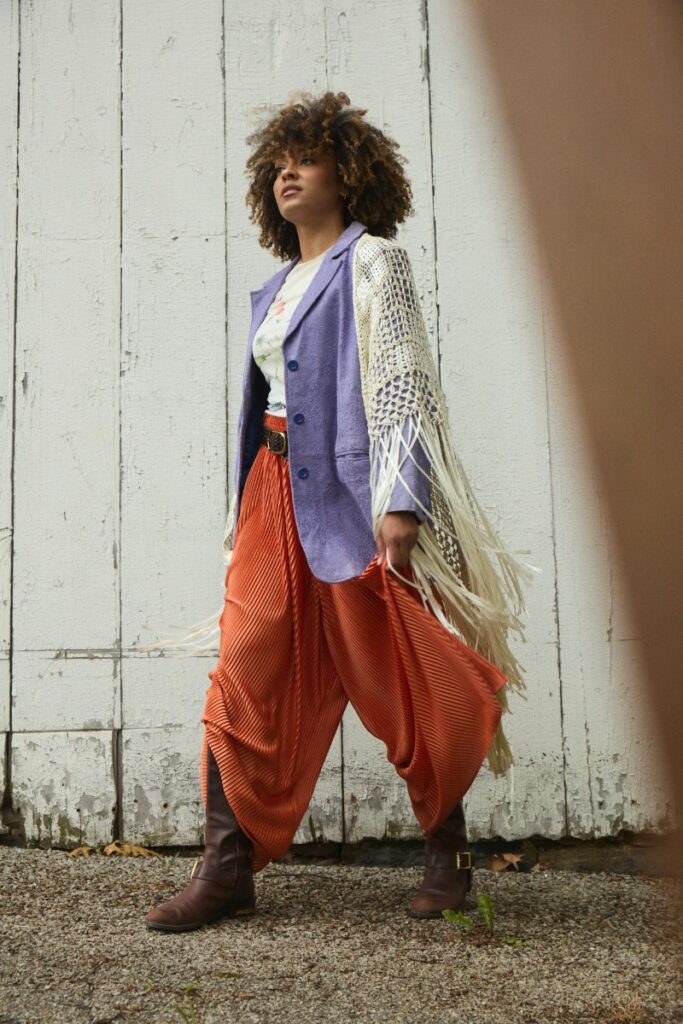 A woman in a periwinkle blazer with a fringed crochet shawl, orange pleated trousers, and knee-high leather boots posing by a white wooden wall.